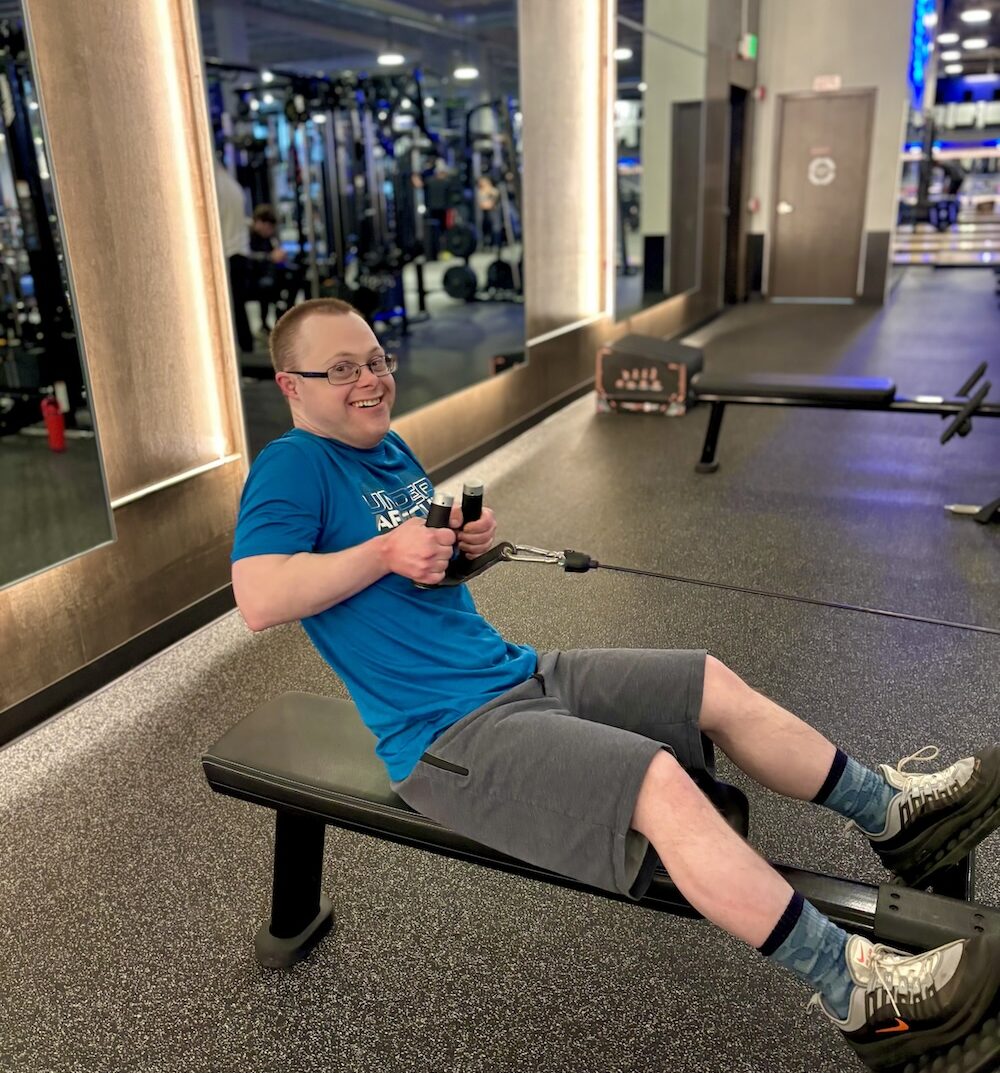 A smiling gym member sits on a bench doing a seated cable row. The gym member is surrounded by sleek lighting, mirrors, and strength equipment in the background.